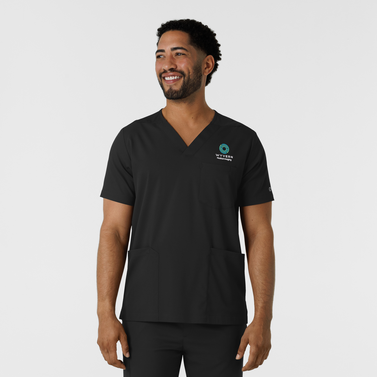 A man wearing a black V-neck scrub top with a teal and white 'VITALITY' logo on the left chest, standing against a light gray studio background.