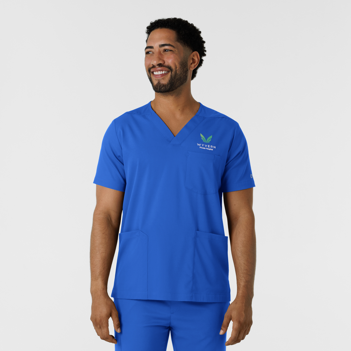 A man wearing a bright blue V-neck medical scrub top and matching scrub pants, standing against a light gray studio background. The scrub top has a small white and green logo on the left chest that reads wyvern health