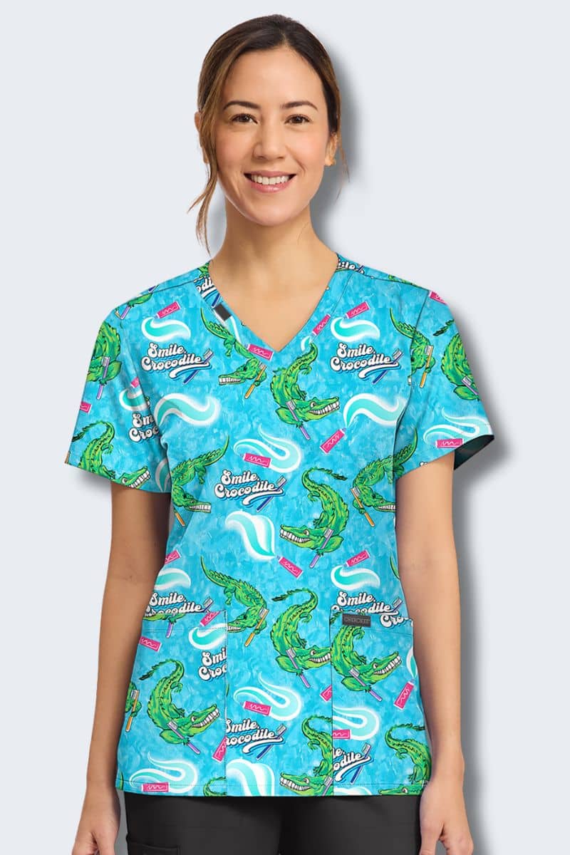 Woman wearing a blue scrub top with alligator pattern on a light gray background. Cherokee Print scrubs CK856_SLCI
