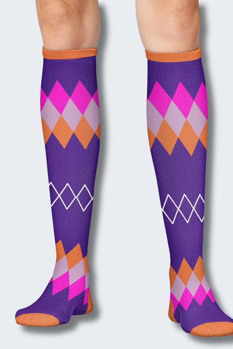A pair of knee-high argyle socks with a purple base, featuring pink, orange, light gray, and white diamond patterns, modeled on legs against a light gray background. Cherokee Printsupport compression socks